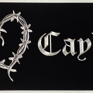 May include: Black license plate featuring a silver barbed wire heart and the name "Cayley" in a silver gothic font.