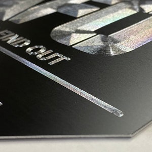 Pu&ograve; includere: Primo piano di un cartello nero con lettere argentate che recitano "AND FIND OUT" e "MORE". Le lettere hanno una finitura testurizzata e iridescente.
