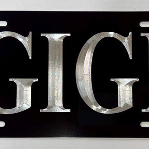 May include: Black license plate with the name "GIGI" in large, silver, block letters. The plate has a glossy finish and includes the standard mounting slots.