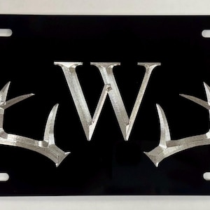 Könnte beinhalten: Schwarzes Nummernschild aus Metall mit einem silbernen 'W'-Monogramm und zwei silbernen Hirschgeweihen auf jeder Seite.