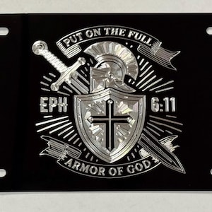 May include: A rectangular black license plate featuring a silver metallic graphic of a helmet, a shield with a cross, and a sword. The text reads "PUT ON THE FULL ARMOR OF GOD" and "EPH 6:11". The plate has four mounting holes, one in each corner.