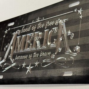 K&ouml;nnte beinhalten: Ein rechteckiges Metall-Nummernschild mit einem schwarz-grauen amerikanischen Flaggendesign im Hintergrund. Der silberne Pr&auml;getext lautet "land of the free", "AMERICA" und "because of the brave", verziert mit silbernen Sternen und Schn&ouml;rkeln.
