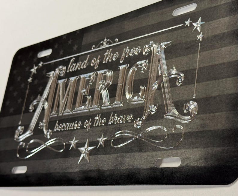 K&ouml;nnte beinhalten: Ein rechteckiges Metall-Nummernschild mit einem schwarz-grauen amerikanischen Flaggendesign im Hintergrund. Der silberne Pr&auml;getext lautet "land of the free", "AMERICA" und "because of the brave", verziert mit silbernen Sternen und Schn&ouml;rkeln.