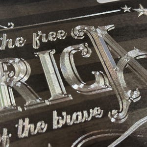 K&ouml;nnte beinhalten: Eine dunkle Holzoberfl&auml;che mit tief eingravierten, metallisch wirkenden Schriftz&uuml;gen, die "of the free" und "of the brave" sowie die gro&szlig;en Buchstaben "RICA" zeigen. Das Design enth&auml;lt dekorative Schn&ouml;rkel und kleine Sternsymbole.