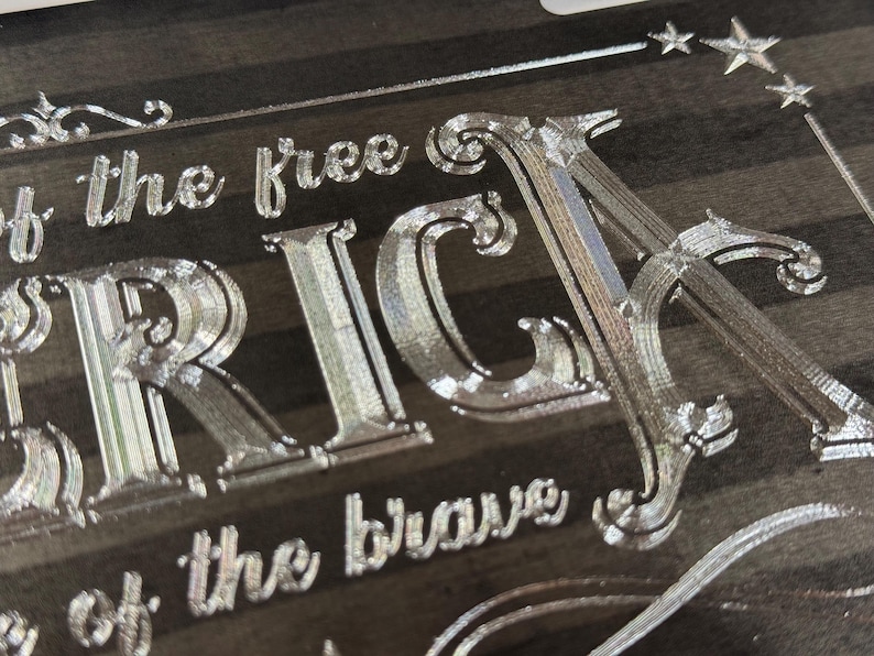 K&ouml;nnte beinhalten: Eine dunkle Holzoberfl&auml;che mit tief eingravierten, metallisch wirkenden Schriftz&uuml;gen, die "of the free" und "of the brave" sowie die gro&szlig;en Buchstaben "RICA" zeigen. Das Design enth&auml;lt dekorative Schn&ouml;rkel und kleine Sternsymbole.