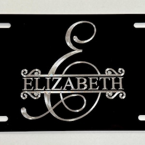 May include: Black license plate with a silver monogram featuring the name "ELIZABETH" and the letter "E". The plate has four mounting holes and a glossy finish.