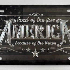 K&ouml;nnte beinhalten: Ein rechteckiges Metall-Nummernschild mit einem abgenutzten schwarz-grauen amerikanischen Flaggendesign. Der silberfarbene Schriftzug lautet "land of the free" &uuml;ber dem gro&szlig;en Wort "AMERICA" und "because of the brave" darunter, verziert mit Sternen.