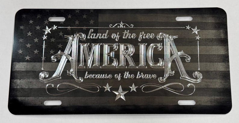 K&ouml;nnte beinhalten: Ein rechteckiges Metall-Nummernschild mit einem abgenutzten schwarz-grauen amerikanischen Flaggendesign. Der silberfarbene Schriftzug lautet "land of the free" &uuml;ber dem gro&szlig;en Wort "AMERICA" und "because of the brave" darunter, verziert mit Sternen.