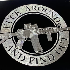 Può includere: Una targa metallica argentata con il testo "F**k Around And Find Out" in un cerchio attorno a una silhouette argentata di un fucile.