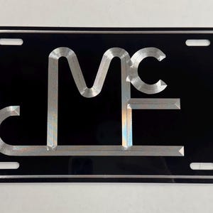 May include: Black license plate with a silver border and the letters "MC" in a stylized, silver font. The plate has four mounting holes and a reflective surface.