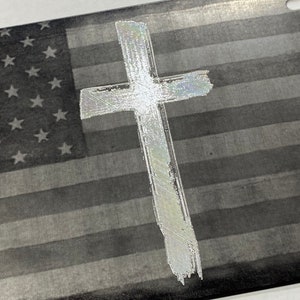 May include: A silver metal license plate with a black and white American flag design and a white cross in the center.