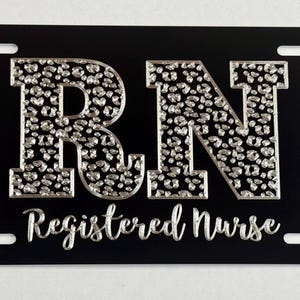 May include: Black license plate featuring silver leopard print letters "RN" and the words "Registered Nurse" in script. The plate has four mounting holes, suitable for a vehicle.
