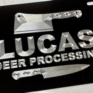May include: A black metal license plate with silver lettering that reads "LUCAS DEER PROCESSING". Two silver knives are displayed on the plate, one is a butcher knife and the other is a smaller knife.