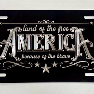 K&ouml;nnte beinhalten: Ein rechteckiges schwarzes Nummernschild mit silberner metallischer Schrift und dekorativen Sternen. Der Text lautet "land of the free" &uuml;ber dem gro&szlig;en Wort "AMERICA" und "because of the brave" darunter, alles eingerahmt von kunstvollen silbernen Verzierungen.