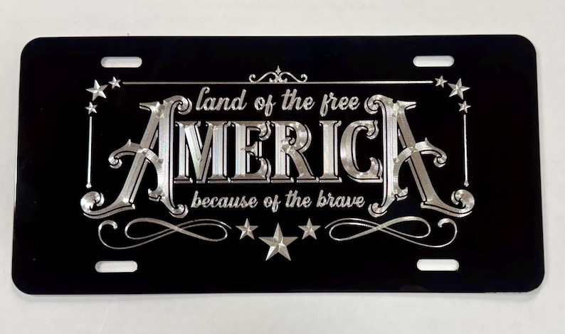 K&ouml;nnte beinhalten: Ein rechteckiges schwarzes Nummernschild mit silberner metallischer Schrift und dekorativen Sternen. Der Text lautet "land of the free" &uuml;ber dem gro&szlig;en Wort "AMERICA" und "because of the brave" darunter, alles eingerahmt von kunstvollen silbernen Verzierungen.