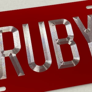Peut inclure: Une plaque d'immatriculation en métal rouge avec le nom "RUBY" en relief en lettres argentées.