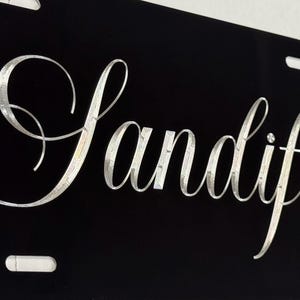 May include: Black license plate with the name "Sandifer" in elegant, silver script. The script is in a flowing, cursive font. The plate has four rectangular slots for mounting.
