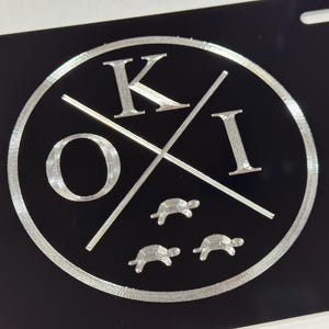 May include: A black and silver circular design with the letters "K" and "I" crossed over the letters "O" and "I". Three silver turtle silhouettes are below the letters.