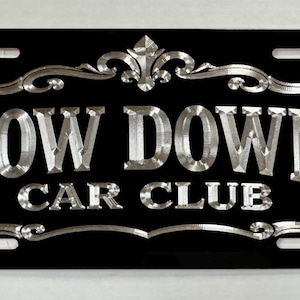 May include: A rectangular black license plate frame featuring silver-colored, metallic-finish lettering that reads LOW DOWN CAR CLUB. The text is enclosed within an ornate, decorative silver border with a fleur-de-lis accent at the top center.