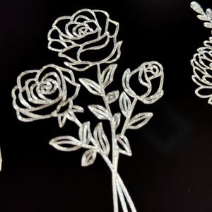 Peut inclure: Un bouquet de trois roses avec des tiges et des feuilles, dessin&eacute; en paillettes argent&eacute;es sur un fond noir.