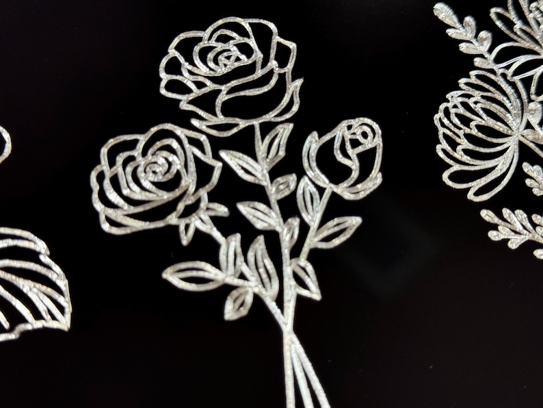 Peut inclure: Un bouquet de trois roses avec des tiges et des feuilles, dessin&eacute; en paillettes argent&eacute;es sur un fond noir.