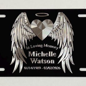 May include: Black license plate with a silver heart, angel wings, and a halo design. The text reads "In Loving Memory Michelle Watson 01/14/1989 - 03/02/2026". The plate has four mounting holes.