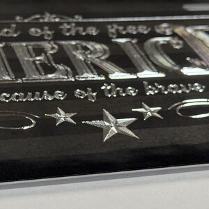 K&ouml;nnte beinhalten: Ein schwarzes Nummernschild mit einem metallisch silbernen amerikanischen Flaggendesign, dem Text "land of the free" &uuml;ber dem Wort "AMERICA" und "because of the brave" darunter, akzentuiert durch drei silberne Sterne und dekorative Schn&ouml;rkel.
