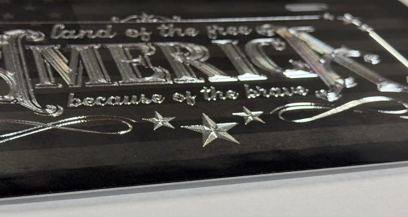 K&ouml;nnte beinhalten: Ein schwarzes Nummernschild mit einem metallisch silbernen amerikanischen Flaggendesign, dem Text "land of the free" &uuml;ber dem Wort "AMERICA" und "because of the brave" darunter, akzentuiert durch drei silberne Sterne und dekorative Schn&ouml;rkel.