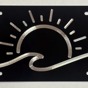 Può includere: Una targa rettangolare nera con un design metallico argentato ritagliato raffigurante un sole nascente sopra un'onda oceanica stilizzata. La targa include quattro fessure di montaggio, due nella parte superiore e due in quella inferiore.