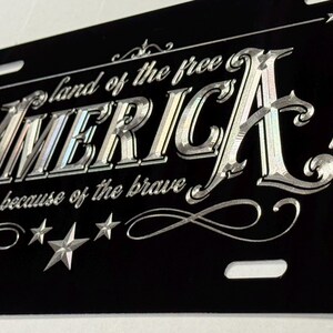 K&ouml;nnte beinhalten: Ein schwarzes, rechteckiges Nummernschild mit silberner Gravur, die den Text "land of the free AMERICA because of the brave" zeigt. Das Design enth&auml;lt dekorative Schn&ouml;rkel und mehrere f&uuml;nfzackige Sterne in den Ecken.