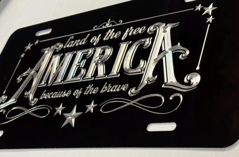 K&ouml;nnte beinhalten: Ein schwarzes, rechteckiges Nummernschild mit silberner Gravur, die den Text "land of the free AMERICA because of the brave" zeigt. Das Design enth&auml;lt dekorative Schn&ouml;rkel und mehrere f&uuml;nfzackige Sterne in den Ecken.