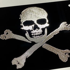 May include: Black license plate featuring a silver skull and crossbones design. The skull has detailed shading, and the bones are crossed beneath it. The design is centered on the plate, creating a bold, pirate-themed aesthetic.