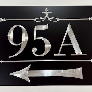 May include: Black metal address sign with the number "95A" in silver. The sign has an arrow pointing left and decorative flourishes. The sign is rectangular with rounded corners and has holes for mounting.