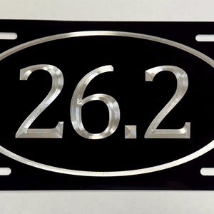 May include: Black license plate with the number "26.2" in a silver oval. The numbers are also silver and are the focal point of the plate. The plate has four mounting holes.