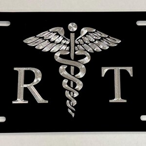 May include: Black license plate with silver-colored medical symbol and the letters "R" and "T". The medical symbol features wings and a staff with a serpent wrapped around it. The letters are in a bold, sans-serif font.