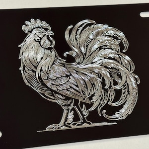 May include: A silver rooster with a detailed design is etched onto a black metal license plate. The rooster is facing right and has its tail feathers spread out.