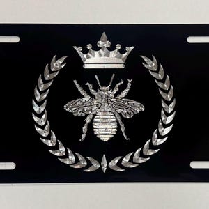 May include: Black license plate featuring a silver bee emblem, a crown, and a laurel wreath design. The bee is centered, with the crown above and the wreath encircling the bee. The plate has four mounting holes.