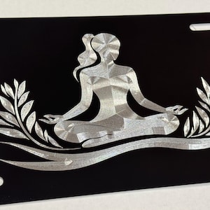 May include: Black license plate featuring a silver illustration of a person in a yoga pose. The figure is surrounded by stylized leaves and sits atop a curved line. The plate has four oblong holes for mounting.