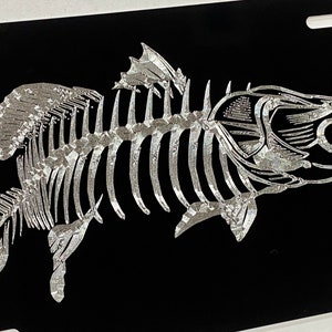 May include: A silver-colored fish skeleton on a black background. The fish is facing right and has a detailed skeletal structure.