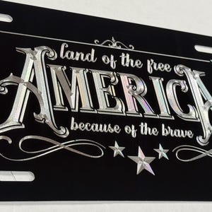 K&ouml;nnte beinhalten: Ein schwarzes, rechteckiges Nummernschild mit silberner, metallischer Schrift, die "land of the free AMERICA because of the brave" lautet. Das Design enth&auml;lt dekorative Schn&ouml;rkel und Sternakzente in den Ecken sowie entlang der unteren Kante.