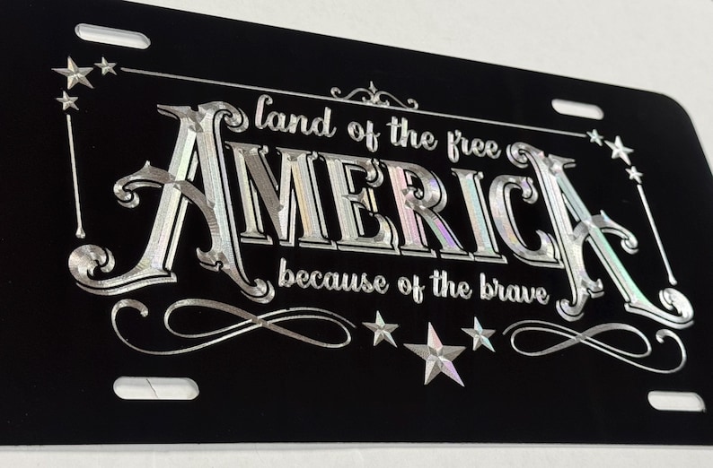 K&ouml;nnte beinhalten: Ein schwarzes, rechteckiges Nummernschild mit silberner, metallischer Schrift, die "land of the free AMERICA because of the brave" lautet. Das Design enth&auml;lt dekorative Schn&ouml;rkel und Sternakzente in den Ecken sowie entlang der unteren Kante.