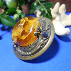 May include: A vintage brooch with a large amber stone set in a gold-toned metal frame. The frame is decorated with intricate floral designs and a small black stone.