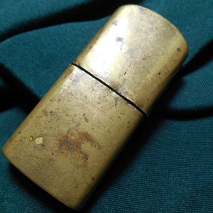 Puede incluir: Un encendedor vintage de latón con un acabado desgastado. El encendedor está cerrado y tiene una forma rectangular.