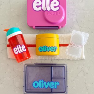 Könnte beinhalten: Eine Sammlung von Lunch-Zubehör, darunter eine rosa Lunchbox mit dem Text "elle", eine rote Wasserflasche mit dem Text "elle", ein gelber Behälter mit dem Text "oliver" und eine lila Lunchbox mit dem Text "oliver".