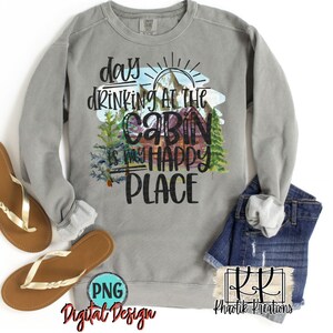 May include: A grey sweatshirt with the text "day drinking at the cabin is my happy place" in black lettering. The text is surrounded by a watercolour illustration of a cabin in the woods with a sun and mountains in the background.