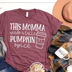Puede incluir: Una camiseta color marrón con el texto "This momma needs a latte pumpkin spice" y un gráfico blanco de una taza de café con un diseño de remolino.