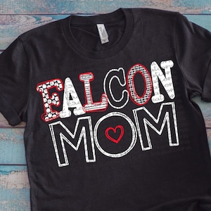 以下が含まれることがあります： 赤いアクセントと赤いハートで白い「FALCON MOM」の文字が書かれた黒いTシャツ。Tシャツは青い木製の背景に置かれ、赤いコンバースの靴と青いデニムのショートパンツが置かれている。