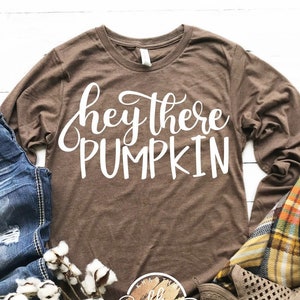 K&ouml;nnte beinhalten: Ein braunes Langarmshirt mit dem Text "hey there PUMPKIN" in wei&szlig;en Buchstaben. Das Shirt wird mit einer blauen Jeans, einer karierten Decke und braunen Cut-Out-Stiefeletten kombiniert.