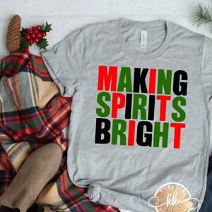 May include: A gray t-shirt with the text "MAKING SPIRITS BRIGHT" in red, green, and black letters. The shirt is folded and laid on a white surface with a plaid blanket, jeans, and pine branches.