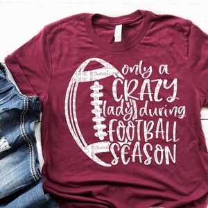 Puede incluir: Una camiseta color burdeos con un gráfico blanco de un balón de fútbol y el texto "only a crazy lady during football season".